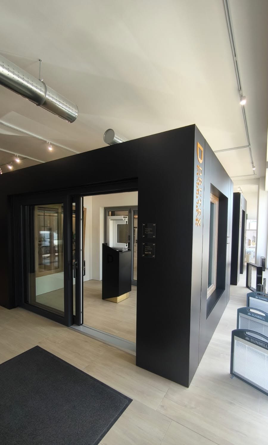 Porta scorrevole vetrata con telaio nero aperta su un cubo espositivo in showroom, a destra espositori Sunbell